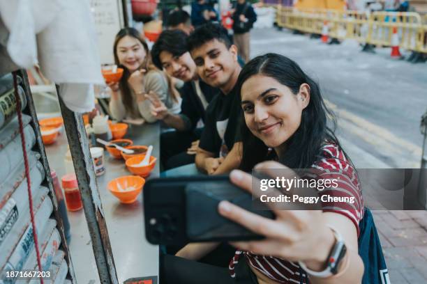 eine junge indische frau macht ein selfie-foto von ihrer freundesgruppe gemischter nationalitäten, während sie zusammen hongkonger streetfood essen - kleinere sehenswürdigkeit stock-fotos und bilder