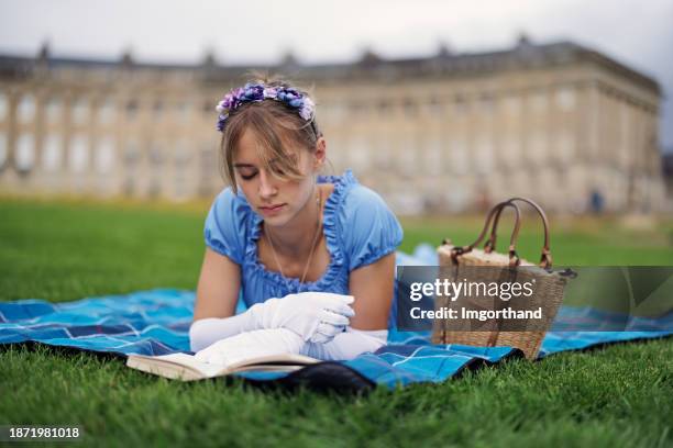 young woman wearing a regency era dress is reading a book in a public park in front of royal crescent - girl in blue dress stock pictures, royalty-free photos & images