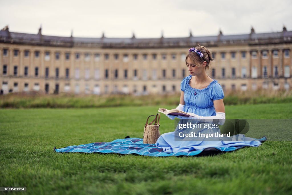 Young woman wearing a regency era dress is reading a book in a public park in front of Royal Crescent