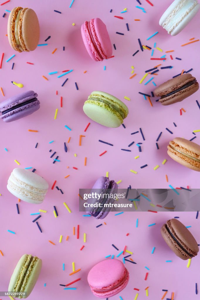 Full frame image of group of multi coloured macarons filled with flavoured butter cream standing on edge, strawberry, vanilla, blueberry, orange, pistachio, chocolate flavoured meringues, spilled sugar sprinkles, purple background, elevated view