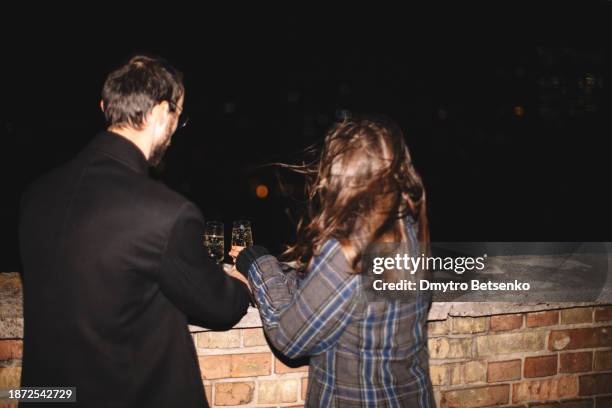 rear view of couple holding champagne flutes during party on the urban rooftop at night - young couple date night wine stock pictures, royalty-free photos & images