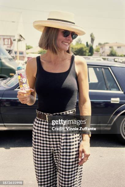 American actress Lynn Herring wearing a black scoop neck top with black-and-white gingham trousers, sunglasses and a white hat, holding a 'Baby...
