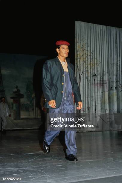 Fashion model walks the runway, wearing blue dungarees beneath a teal jacket with a red beret, an outfit designed by French fashion designer Jean...