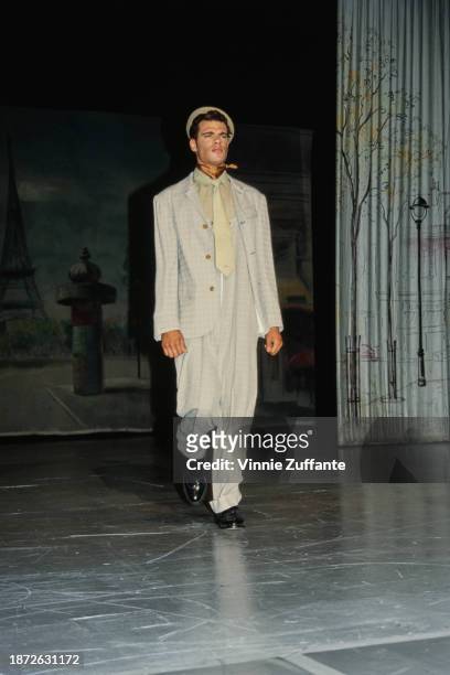 Fashion model walks the runway, wearing a light grey checked suit with high-waisted trousers and a pale green shirt and tie with a bronze...