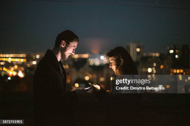 happy young man and young woman using smart phones while standing on the urban rooftop against night city - telephone number stock pictures, royalty-free photos & images