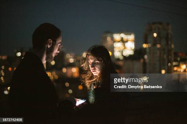 young couple exchanging their phone numbers using smart phones while standing against night city - telephone number stock pictures, royalty-free photos & images