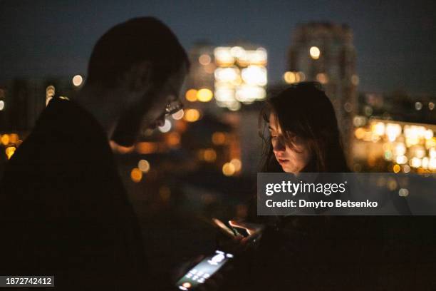 young couple exchanging their phone numbers using smart phones while standing against night city - telephone number stock pictures, royalty-free photos & images