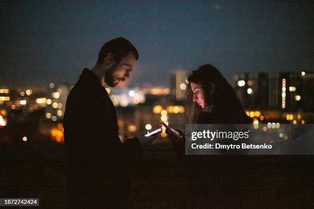 young man and young woman using their smart phones while standing on the urban rooftop against night city - telephone number stock pictures, royalty-free photos & images