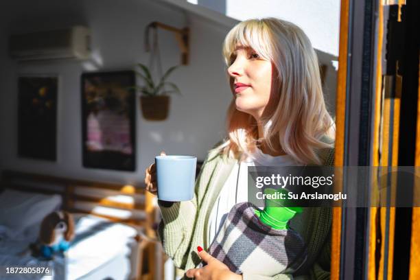 bild einer jungen frau, die aus dem fenster schaut, während sie eine wärmflasche benutzt und heißen tee trinkt - wärmflasche stock-fotos und bilder