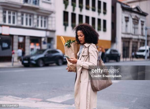 connected errands: smiling woman juggling groceries and phone on the street - baguette stock pictures, royalty-free photos & images
