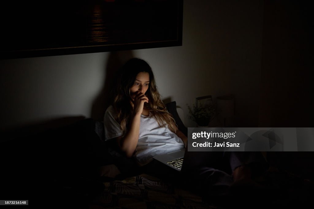 Woman watching a horror movie from her laptop lying on her bed in her bedroom. She has a scary look on her face.