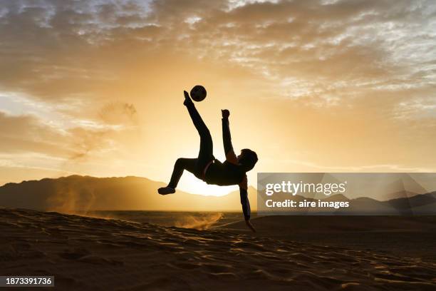 side view of a boy in sportswear jumping in the middle of the desert maneuvering and kicking a soccer ball - children playing silhouette stock pictures, royalty-free photos & images