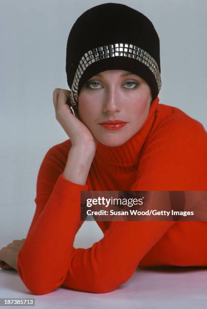 An unidentified model in a red turtleneck sweater and a black cap with appliqued square studs poses against a white background, February 1975. The...