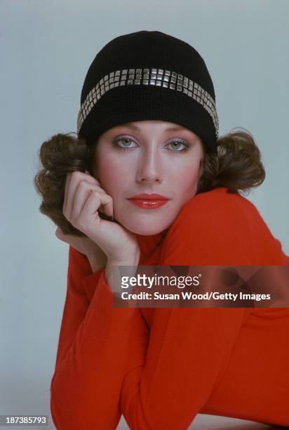 An unidentified model in a red turtleneck sweater and a black cap with appliqued square studs poses against a white background, February 1975. The...