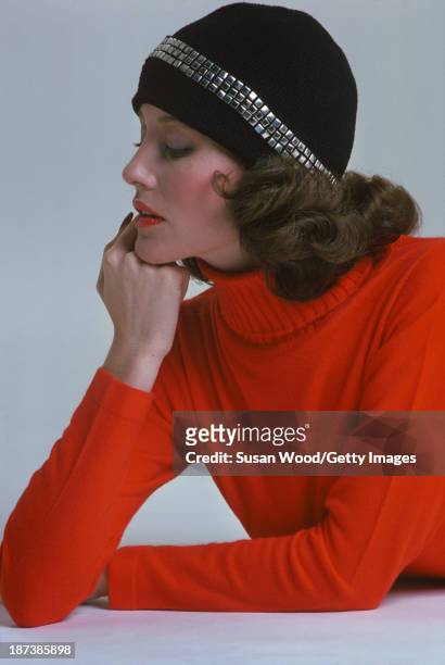 An unidentified model in a red turtleneck sweater and a black cap with appliqued square studs poses against a white background, February 1975. The...