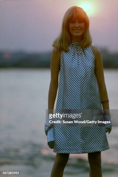 An unidentified model, in a black and white, sleeveless, halter-neck dress, and silver gloves with a silver clutch, poses beside the water at sunset,...