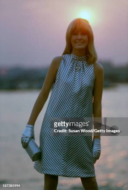 An unidentified model, in a black and white, sleeveless, halter-neck dress, and silver gloves with a silver clutch, poses beside the water at sunset,...