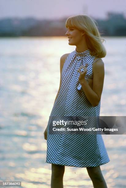 An unidentified model, in a black and white, sleeveless, halter-neck dress and with a silver clutch, poses beside the water at sunset, Malibu,...