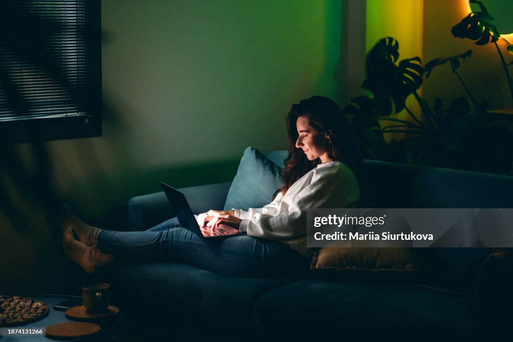 Young adult female using laptop in dark room with cozy ambient lighting.