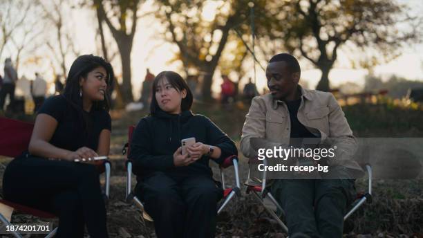 drei multiethnische freunde sitzen auf campingstühlen und unterhalten sich gerne in der natur auf dem campingplatz - wildnisgebiets name stock-fotos und bilder