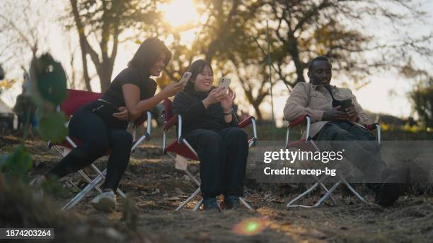 drei multiethnische freunde sitzen auf campingstühlen und unterhalten sich gerne in der natur auf dem campingplatz - wildnisgebiets name stock-fotos und bilder