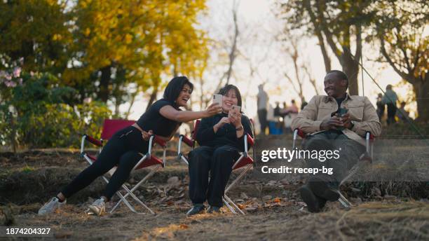 drei multiethnische freunde sitzen auf campingstühlen und unterhalten sich gerne in der natur auf dem campingplatz - wildnisgebiets name stock-fotos und bilder