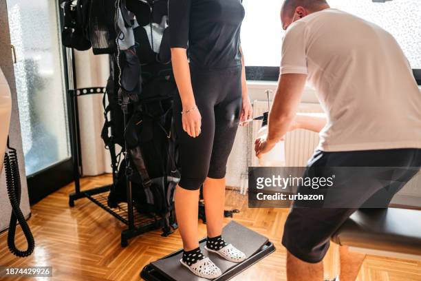 personal trainer hilft seiner kundin, sich im fitnessstudio in einen ems-anzug zu kleiden - ems sport stock-fotos und bilder