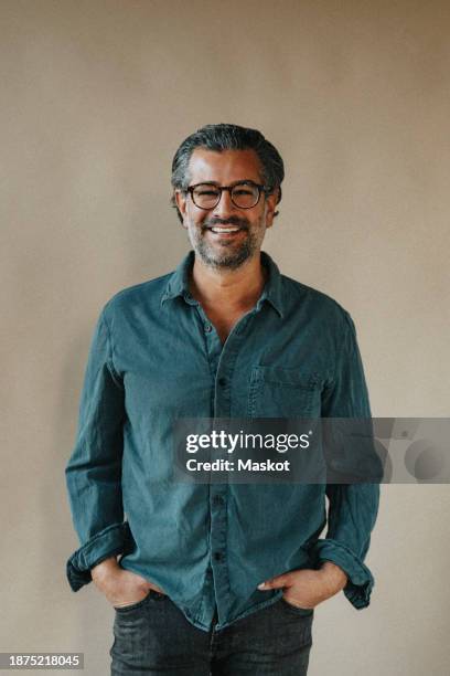 portrait of happy mature man standing with hands in pockets against beige wall - solo un uomo maturo foto e immagini stock