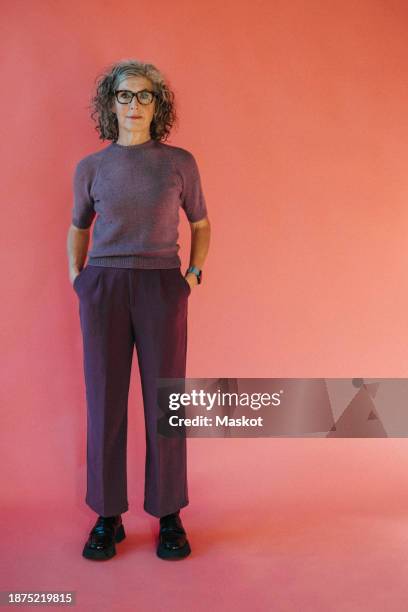 full length portrait of senior woman standing with hands in pockets against red backdrop - figura intera foto e immagini stock