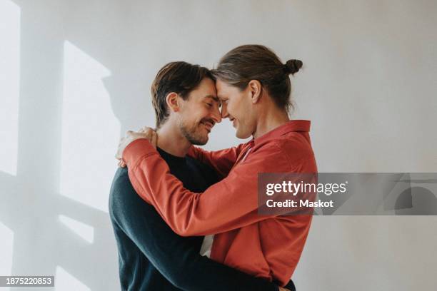 side view of romantic gay couple embracing while standing by white wall - gay man stock pictures, royalty-free photos & images