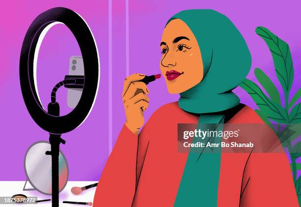 female influencer in hijab giving makeup tutorial with lipstick behind ring light and smart phone - cosmetics stock illustrations