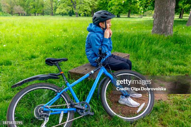 a boy sitting on a park bench with his bicycle next to him - bank müde frühling stock-fotos und bilder