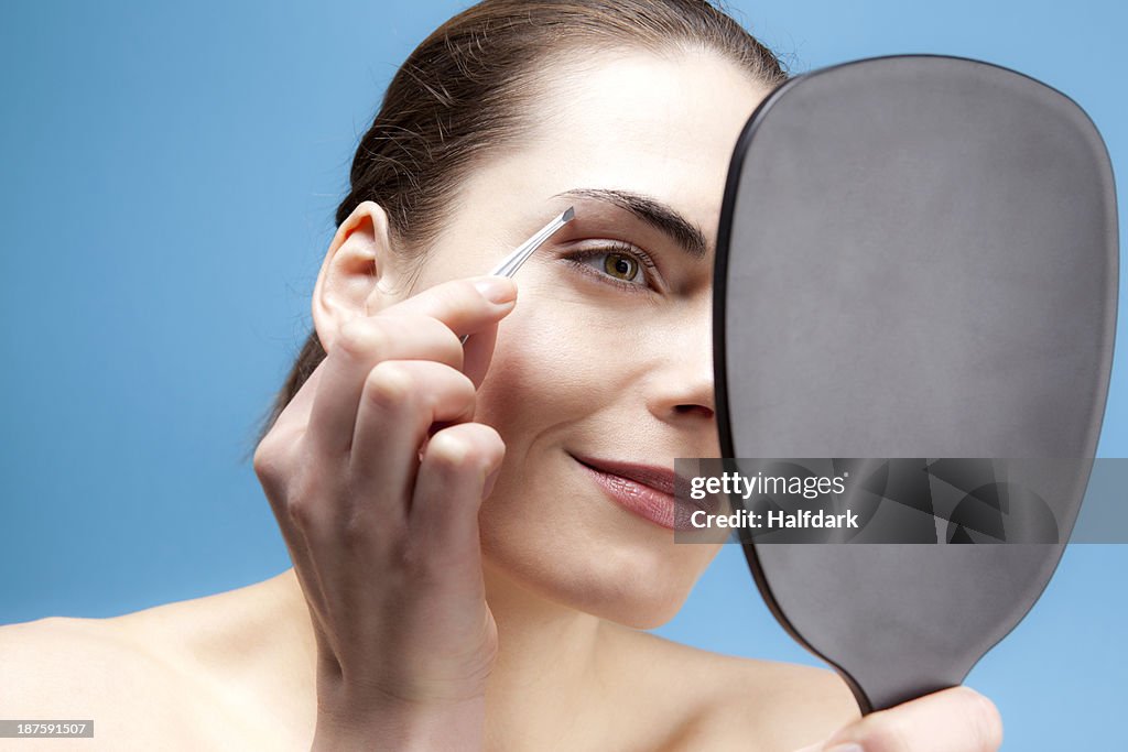A smiling young woman holding a hand mirror and tweezing her brows