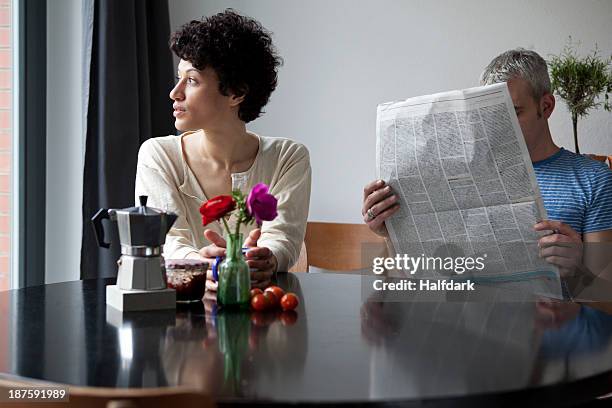 a serious woman looking out a window while her boyfriend hides behind a newspaper - ignore stock-fotos und bilder