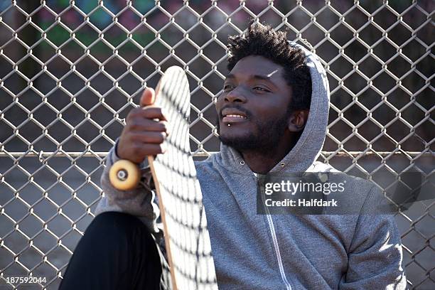 a grinning young man leaning against a chain link fence holding a skateboard - chin piercing stock pictures, royalty-free photos & images