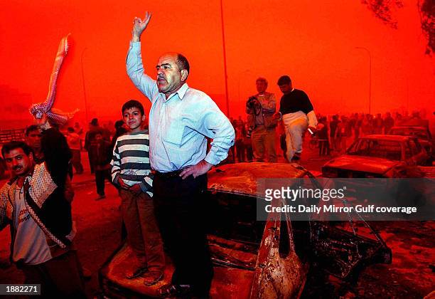 Against a red-tinted sky caused by severe dust storms and burning oil fires, Iraqis protest amid the charred remains of automobiles destroyed in...