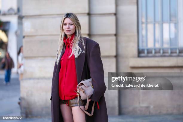 Natalia Verza wears a brown long oversized winter coat, a red sportswear jacket, a beige fluffy bag from Fendi, brown leather mini shorts, during a...