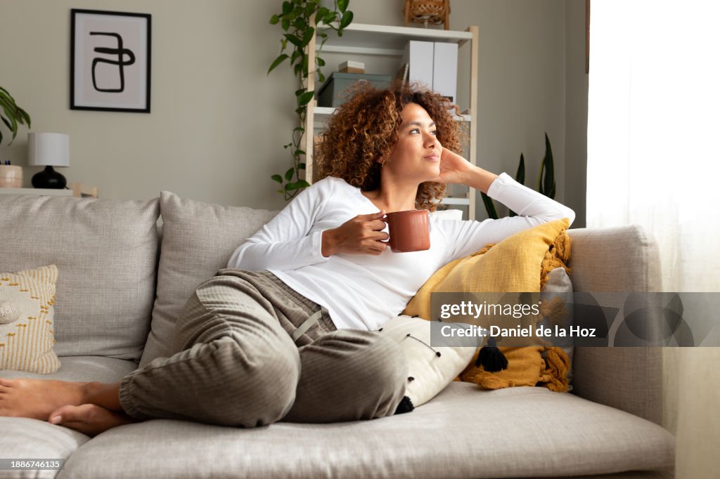Pensive multiracial woman relaxing at home, sitting on the sofa drinking morning coffee.