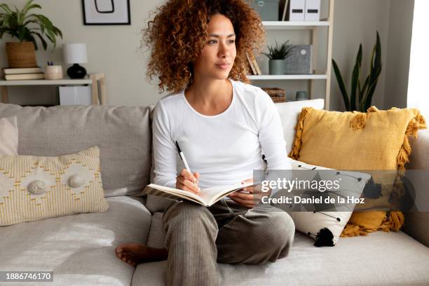 pensive young hispanic woman thinking before writing on personal diary sitting on the couch at home. multiracial latina female writing notes with pen. - écriture manuscrite photos et images de collection