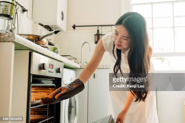 young asian woman wearing protection glove taking food out from oven in kitchen at home. cooking, and people lifestyle concept. - taking off gloves stock pictures, royalty-free photos & images