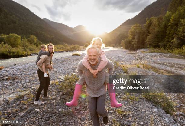 grandfather and father carrying children piggyback at a riverbank at sunset - granddaughter stock pictures, royalty-free photos & images