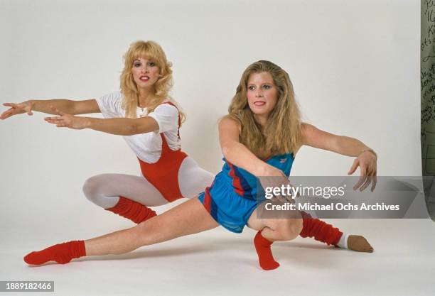Two women modelling exercise wear, one wearing a red leotard over a white t-shirt with light grey leggings and red leg warmers, as the other wears a...