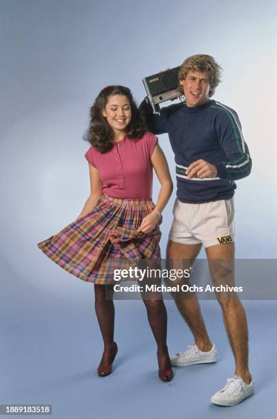 Young woman wearing a pink cap-sleeved top and a pink, yellow and blue plaid skirt, dances beside a young man, who wears a dark blue sweatshirt with...
