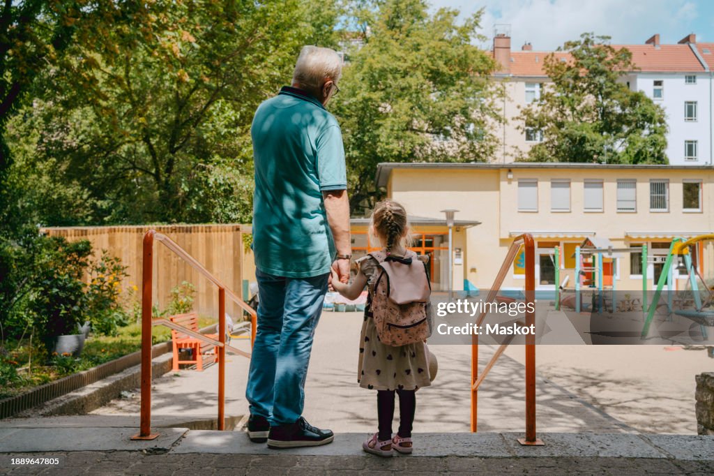 Senior man with granddaughter standing near railing at kindergarten