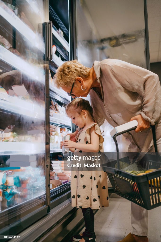 Senior woman with granddaughter choosing frozen food while doing shopping at grocery store