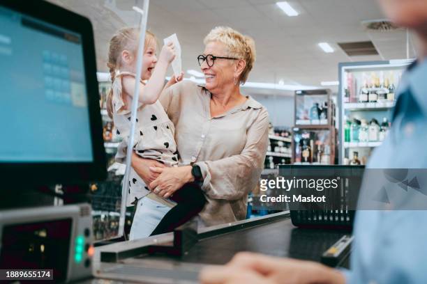 happy senior woman carrying cheerful granddaughter holding bill at supermarket - grandma invoice stock pictures, royalty-free photos & images