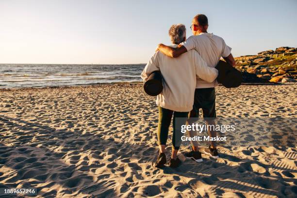 rear view of senior couple holding exercise mats while standing on sand at beach - rolled up yoga mat stock pictures, royalty-free photos & images