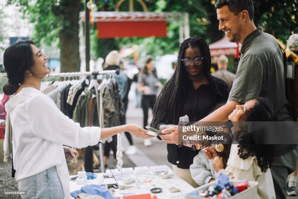 Side view of happy female vendor laughing while taking online payment from customer at flea market