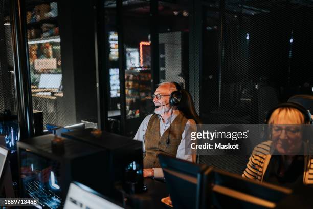 elderly man wearing headset sitting by female friend playing video game on computer at gaming lounge - bonding icon stock pictures, royalty-free photos & images
