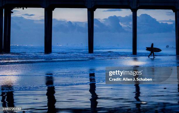 Huntington Beach, CA A surfer exits the surf after riding the last waves of 2023 is silhouetted by cloudy skies at dusk amid cool weather in...